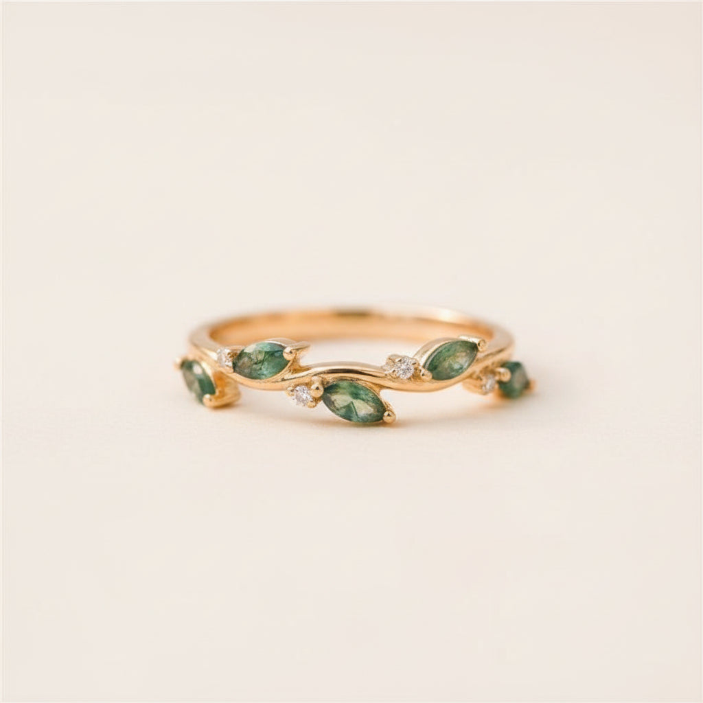A gold ring with marquise-cut green gemstones and small clear accent stones, resting on a textured stone surface with a softly blurred natural background.