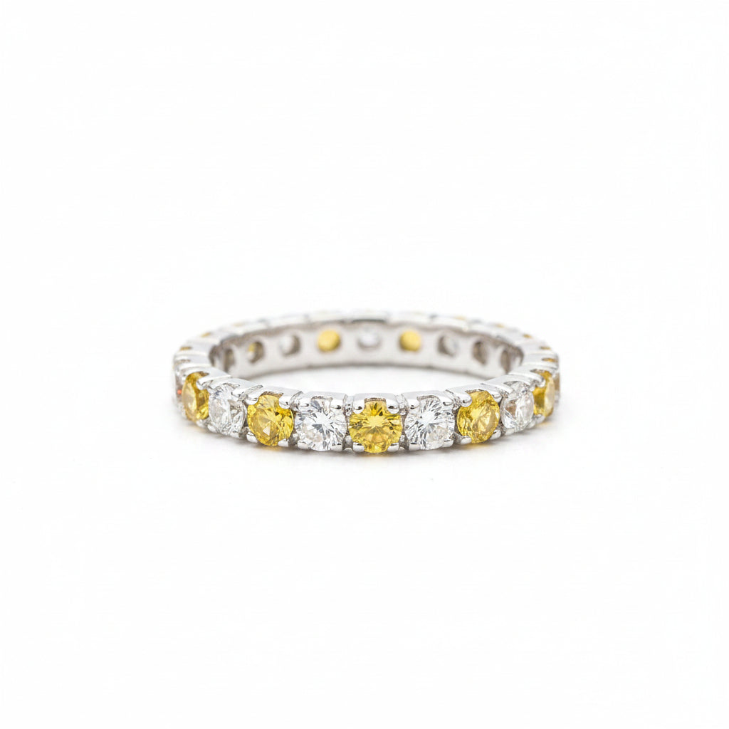 A silver eternity ring featuring alternating yellow gemstone and clear stone segments, resting on a textured light gray stone surface with a blurred green background.
