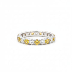 A silver eternity ring featuring alternating yellow gemstone and clear stone segments, resting on a textured light gray stone surface with a blurred green background.