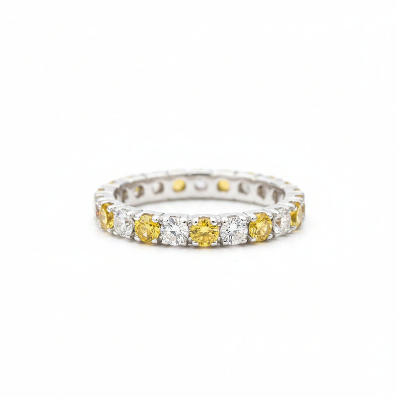 A silver eternity ring featuring alternating yellow gemstone and clear stone segments, resting on a textured light gray stone surface with a blurred green background.