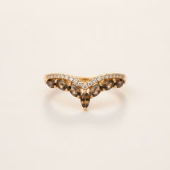 A gold V-shaped ring featuring marquise-cut brown smoky gemstones and a row of small clear stones, resting on a textured stone surface with a blurred green background.