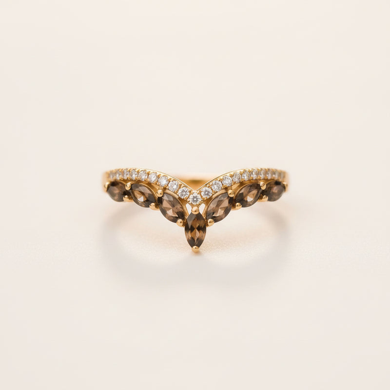 A gold V-shaped ring featuring marquise-cut brown smoky gemstones and a row of small clear stones, resting on a textured stone surface with a blurred green background.