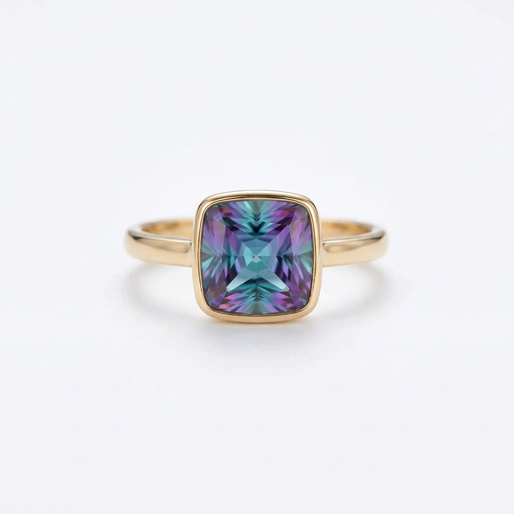 A gold ring with a square-cut iridescent gemstone showing purple, green, and blue colors, resting on a beige lace fabric with dried flowers.