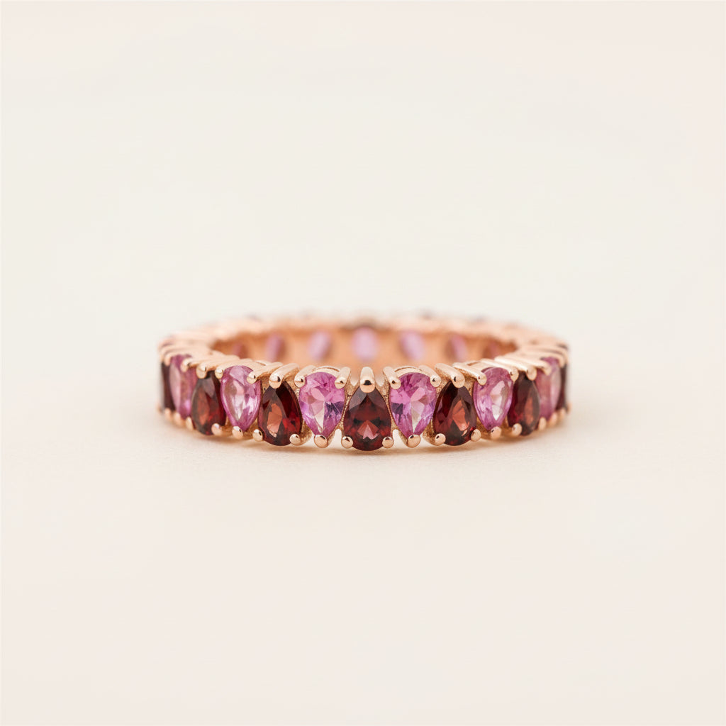 A rose gold eternity ring featuring a row of marquise-cut pink and deep red gemstones, resting on a textured stone surface.