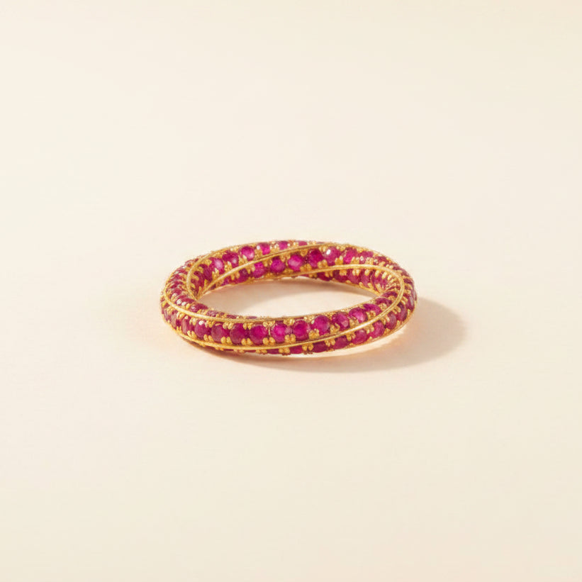 A gold bangle bracelet with rows of round pinkish-red stones, displayed on a clear glass table with a reflection visible below.