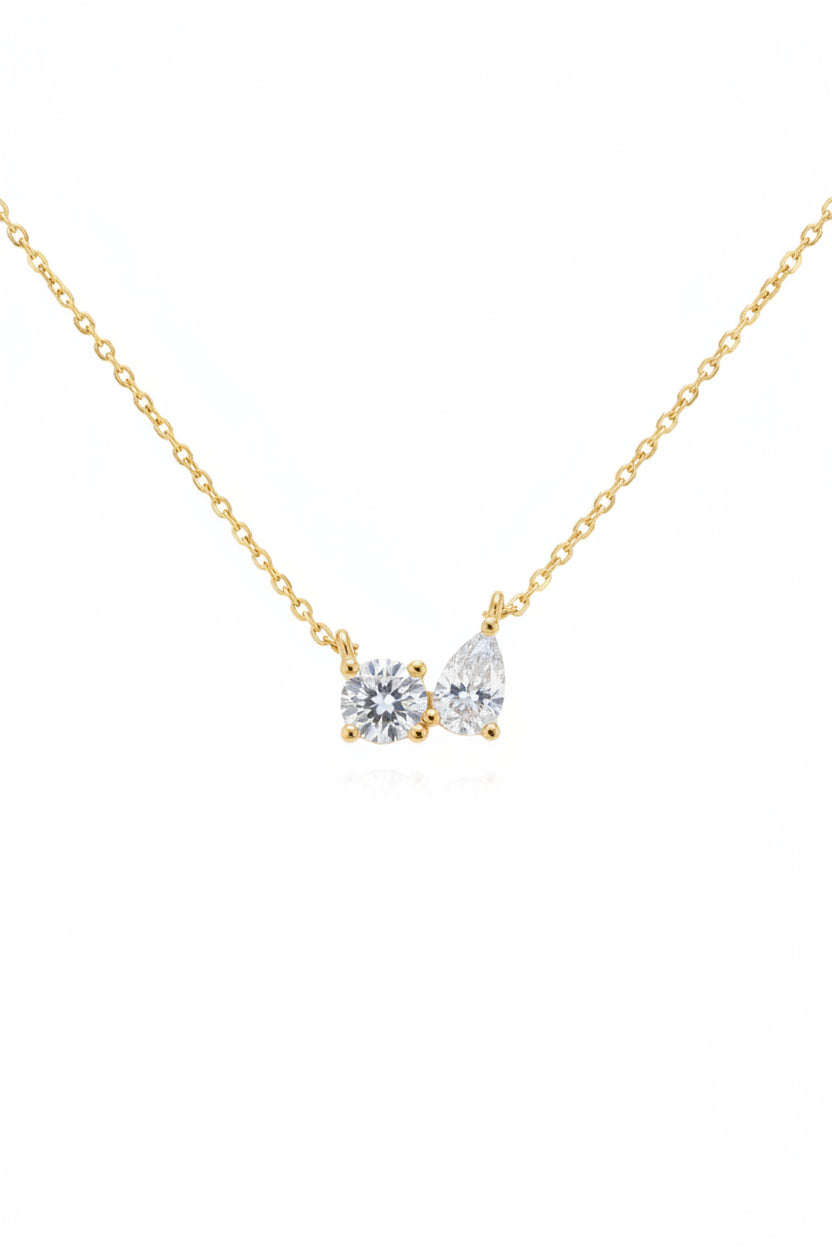A gold chain necklace with a double-pendant design featuring one round clear gemstone and one teardrop-shaped clear gemstone in gold settings, displayed on a dark textured stone surface.