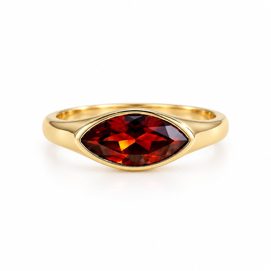 A gold ring with a marquise-cut dark red gemstone, resting on a textured cream lace fabric with soft green leaves blurred in the background.