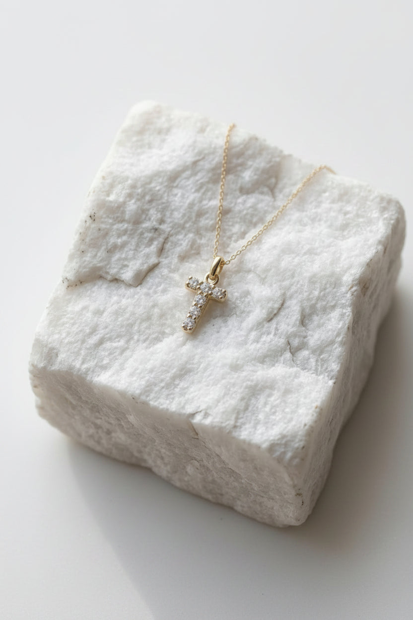 A delicate gold cross pendant necklace adorned with small clear stones, resting on a textured white stone block.