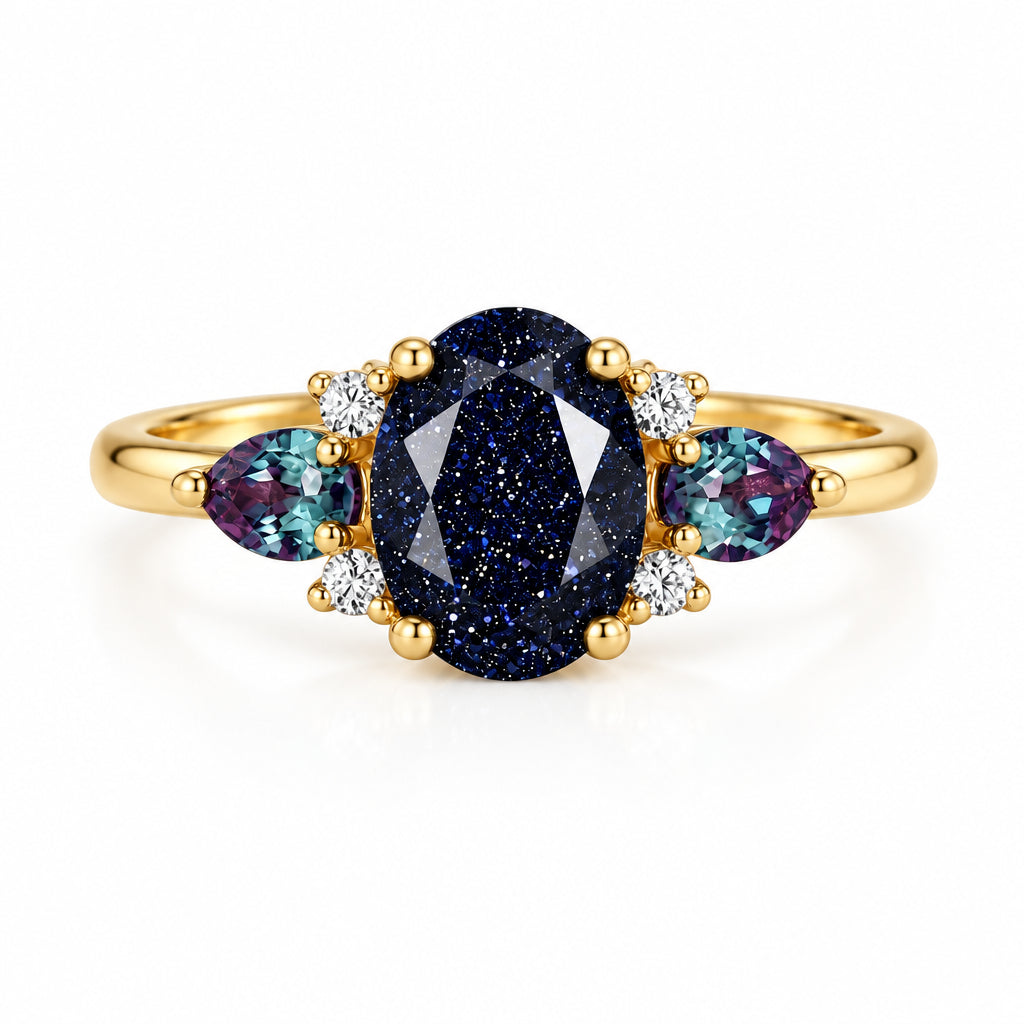 A gold ring featuring a central oval dark blue glitter stone flanked by two marquise-shaped blue stones and small clear accent stones, displayed on a textured beige fabric with soft green leaves in the background.