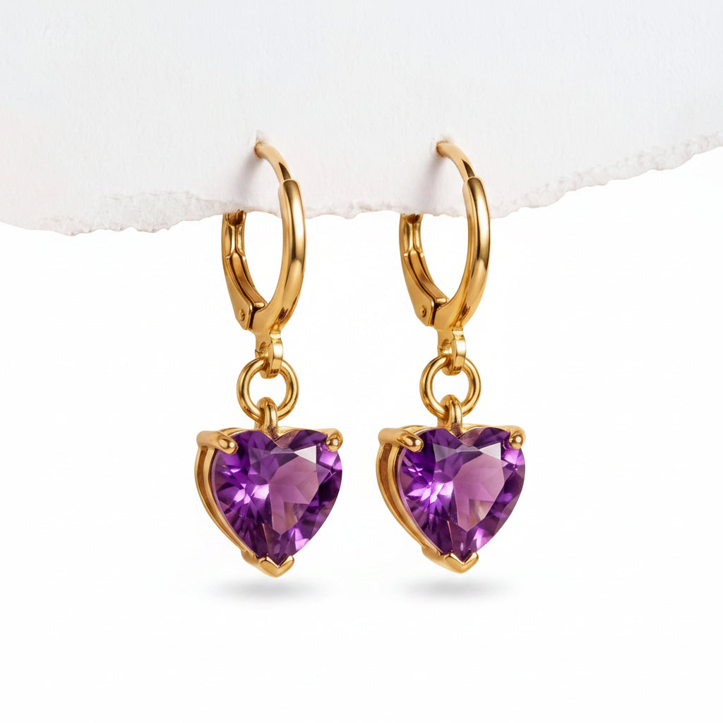 A pair of gold hoop earrings with dangling heart-shaped purple gemstones, displayed against a light beige textured background.