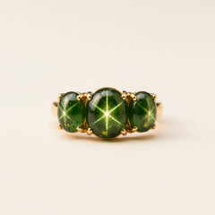 A gold ring featuring three oval green starburst cabochon gemstones, resting on a textured stone surface with a soft-focus natural background.