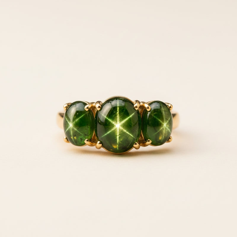 A gold ring featuring three oval green starburst cabochon gemstones, resting on a textured stone surface with a soft-focus natural background.