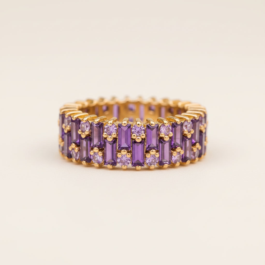 A gold eternity ring set with purple baguette and round-cut gemstones, resting on a textured stone surface with a blurred natural background.
