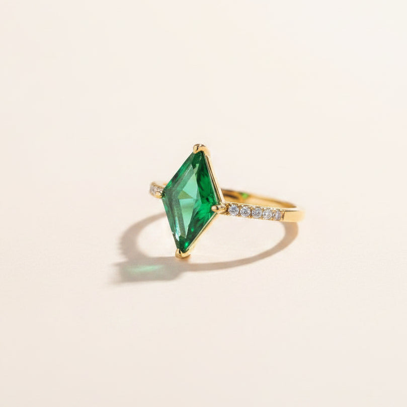 A gold ring with a large emerald-cut green gemstone and small clear accent stones on the band, displayed inside an open navy blue ring box with a white cushion.