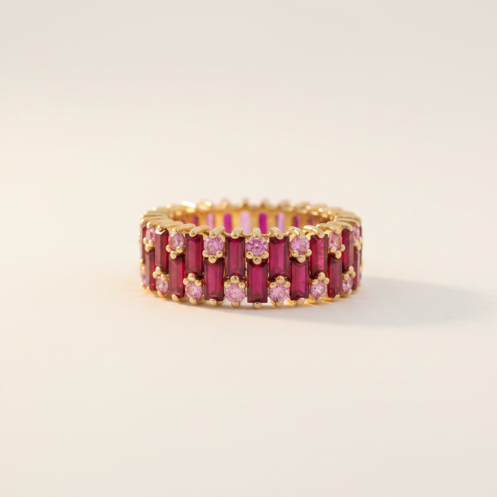 A gold eternity ring featuring rows of rectangular deep pink rubies and small round pink stones, displayed on a white cube against a soft gray background.