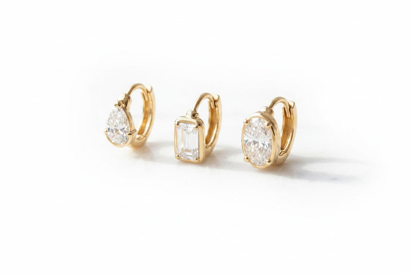 Three gold hoop earrings with clear gemstones in different shapes, displayed on a white marble surface.