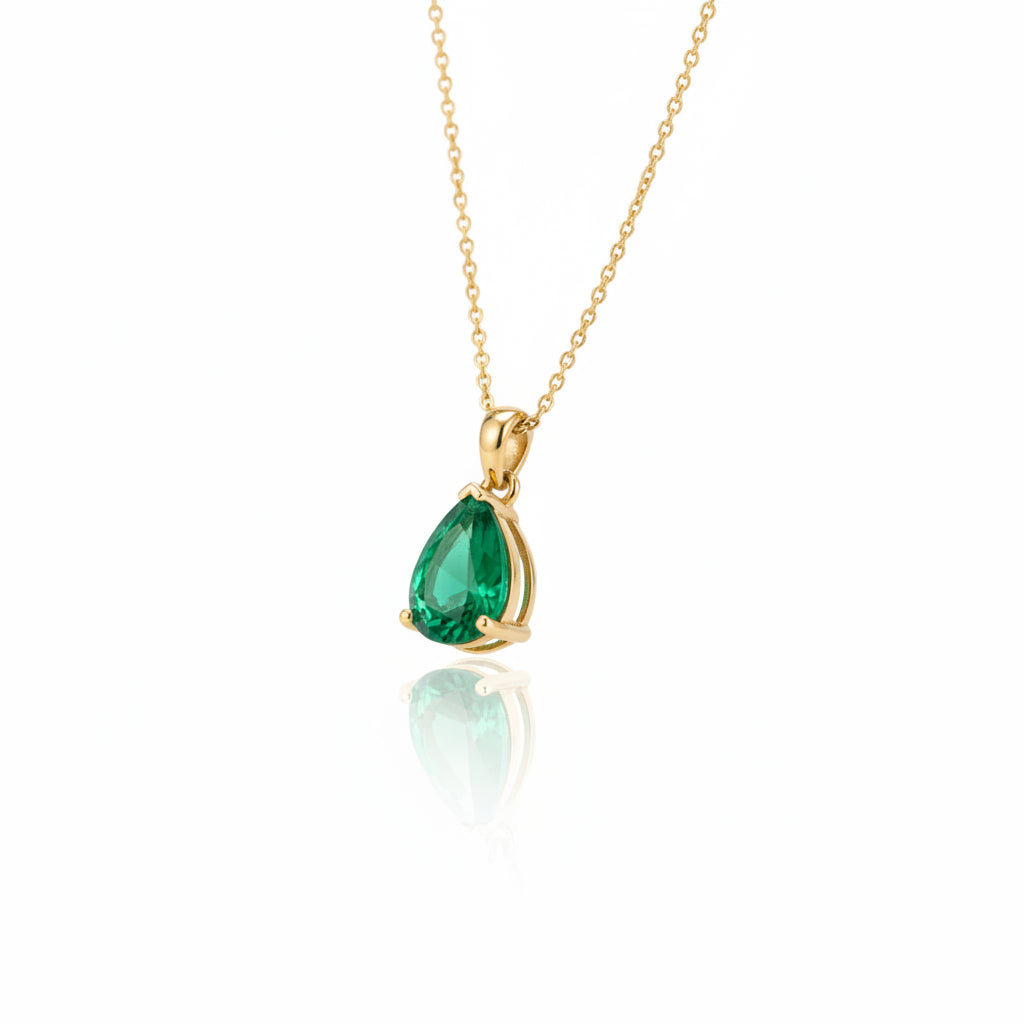 A gold pendant necklace with a teardrop-shaped green gemstone resting on smooth dark river stones with moss in the background.