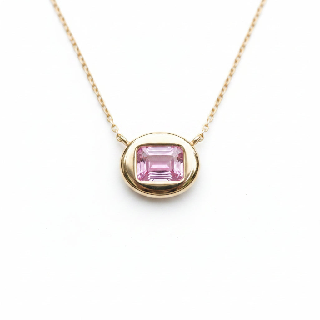 A gold pendant necklace with an emerald-cut pink gemstone, resting on smooth dark green river stones with small green moss visible at the base.