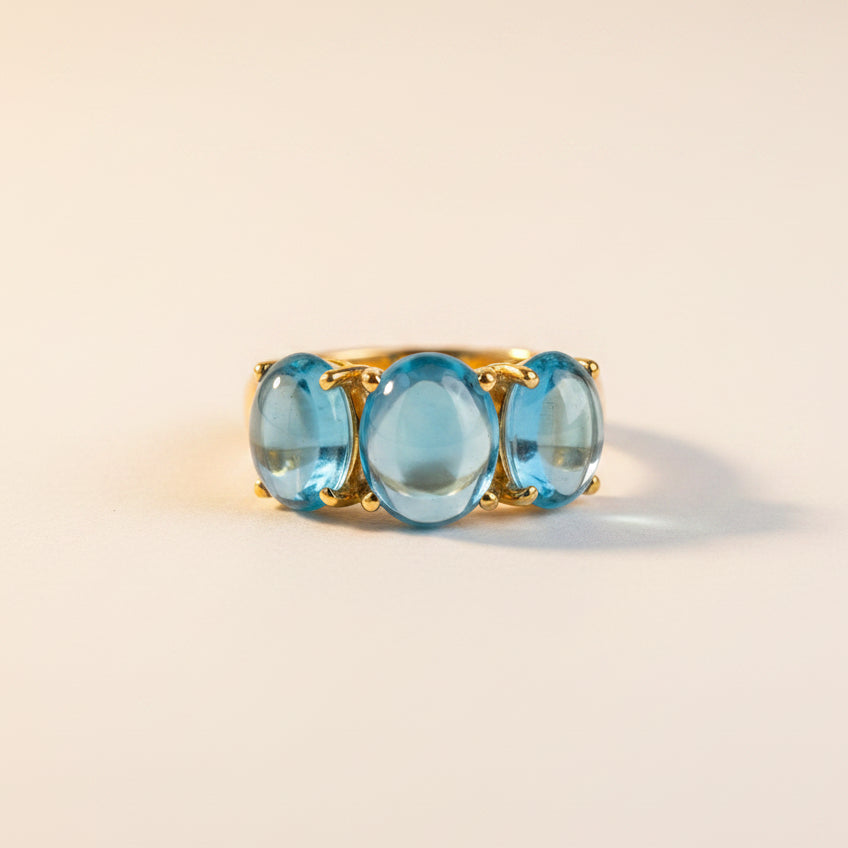 A gold ring with three oval-cut light blue gemstones, presented in an open dark gray velvet jewelry box with a cream interior.