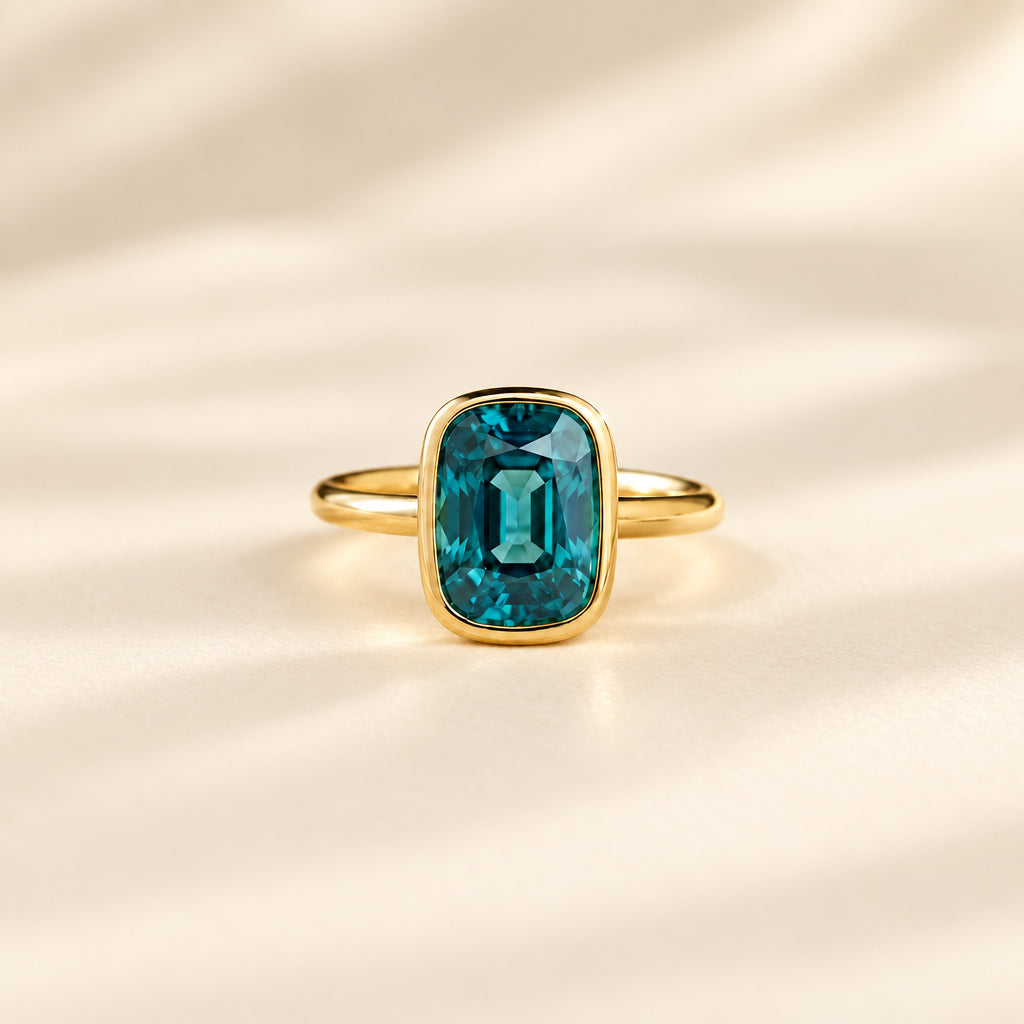 A gold ring with a large rectangular emerald-cut blue-green gemstone, resting on a textured beige lace fabric with dried flowers.