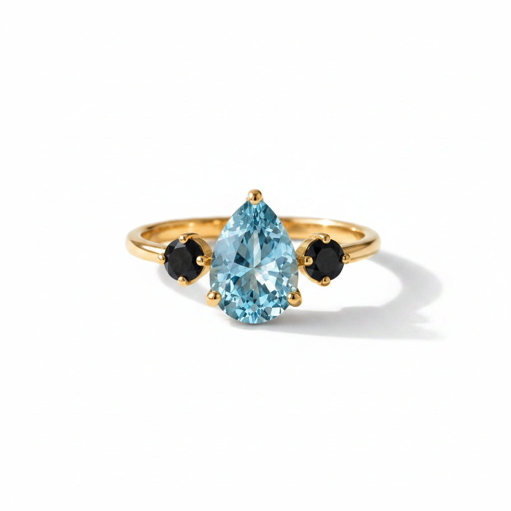 A gold ring featuring a pear-cut light blue center stone and two small round black stones on either side, photographed on a textured beige fabric background with warm sunlight and shadows.