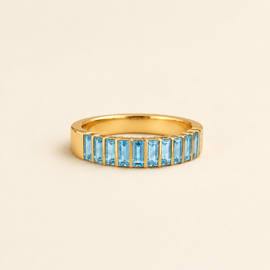 A gold eternity ring featuring a row of rectangular light blue gemstones, resting on a textured beige fabric with soft green leaves blurred in the background.