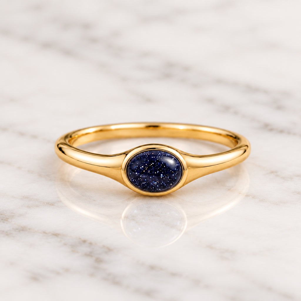 A gold ring with an oval dark blue glitter stone, resting on a textured beige fabric with soft green leaves blurred in the background.