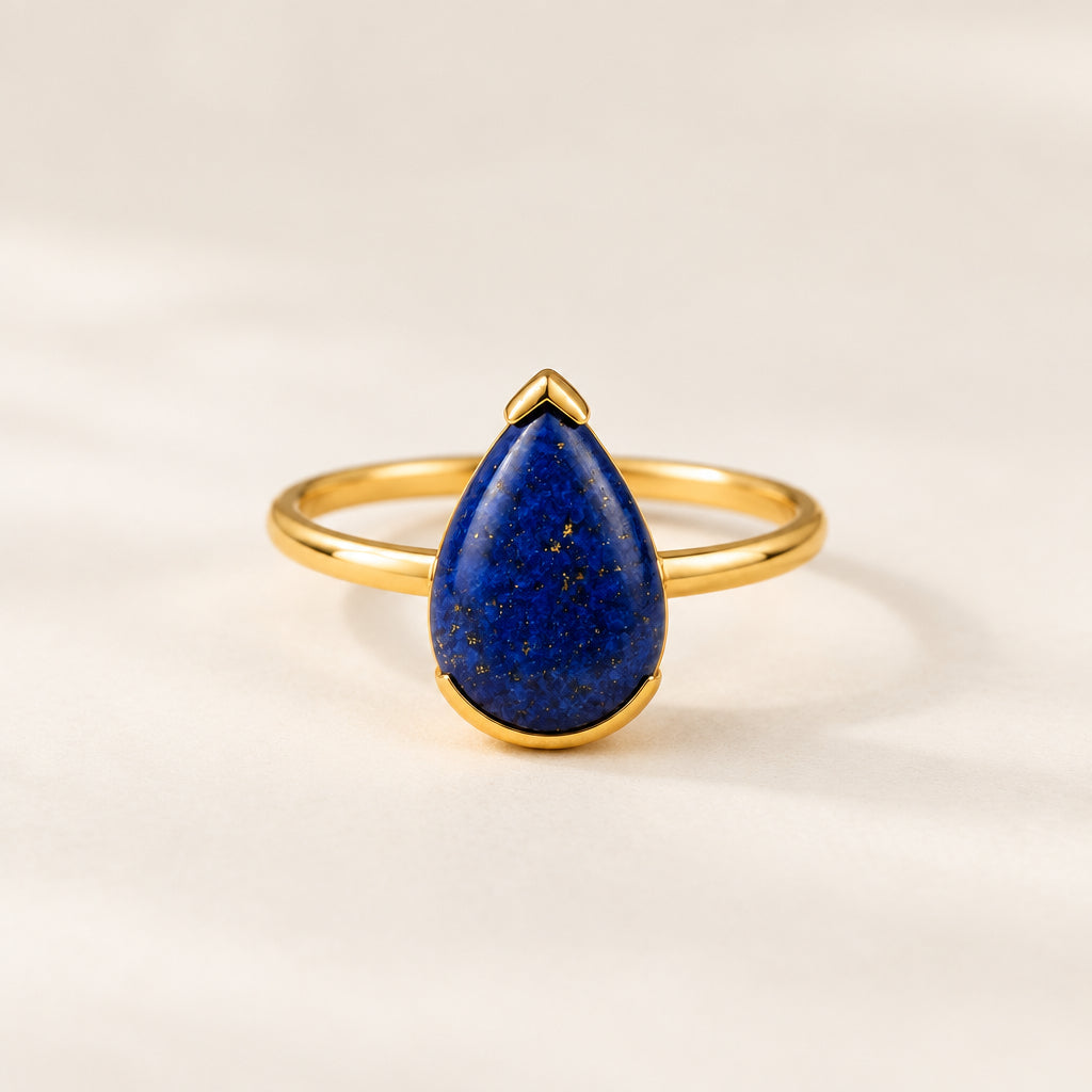 A gold ring with a teardrop-shaped lapis lazuli stone, resting on a textured beige fabric with dried flowers.