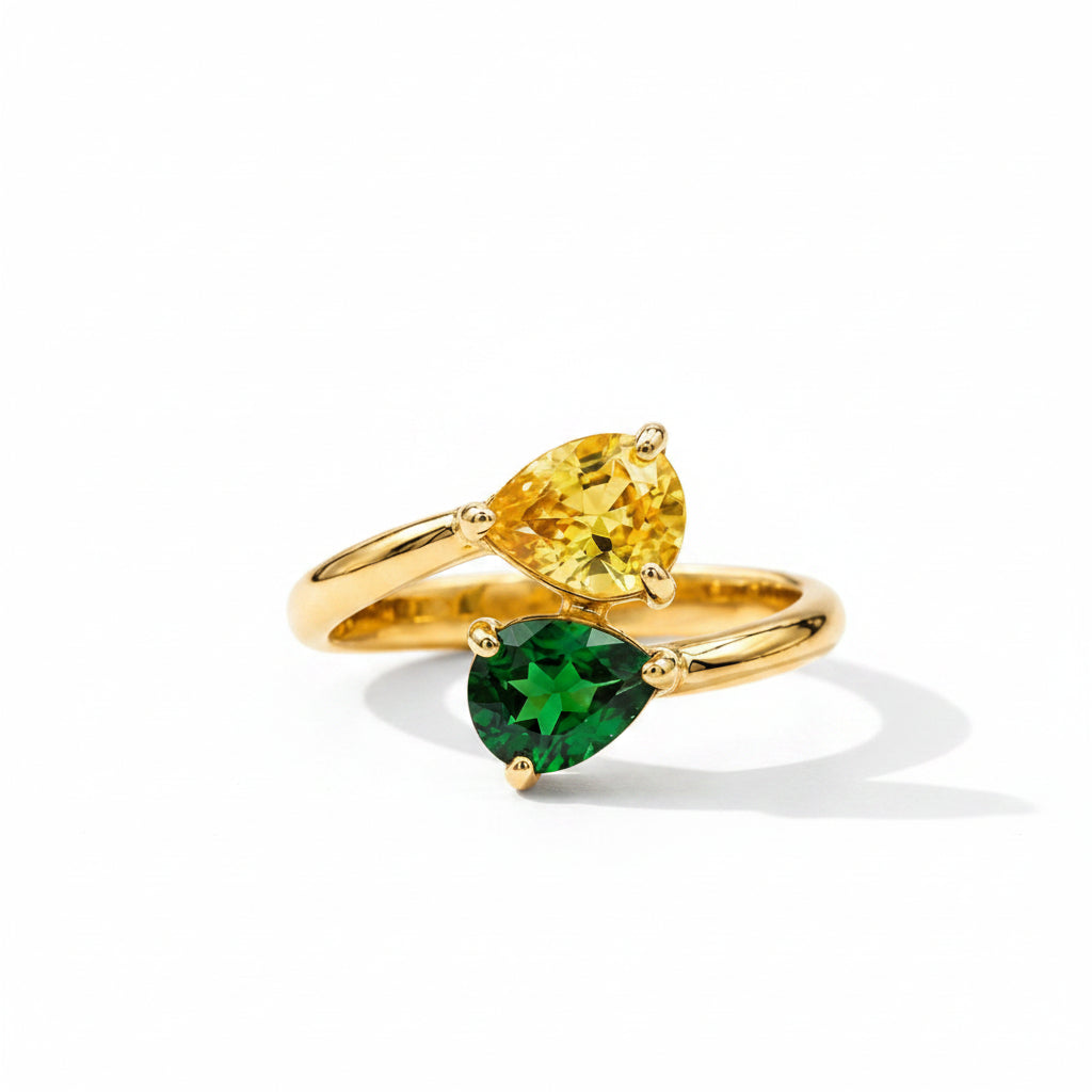 A gold ring set with two pear-cut gemstones, one yellow and one green, displayed on a textured beige fabric background.