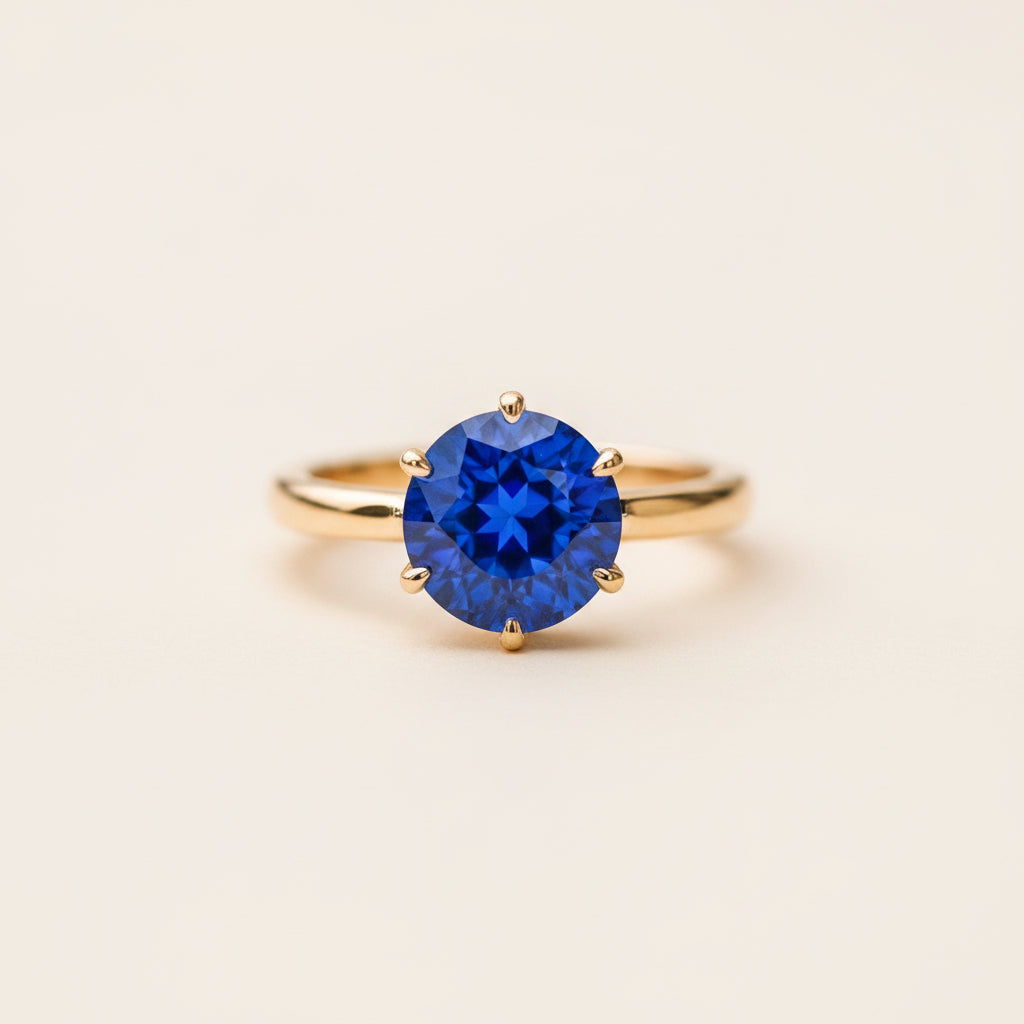 A yellow gold ring with a round brilliant-cut deep blue gemstone, resting on a textured stone surface with a blurred natural background.