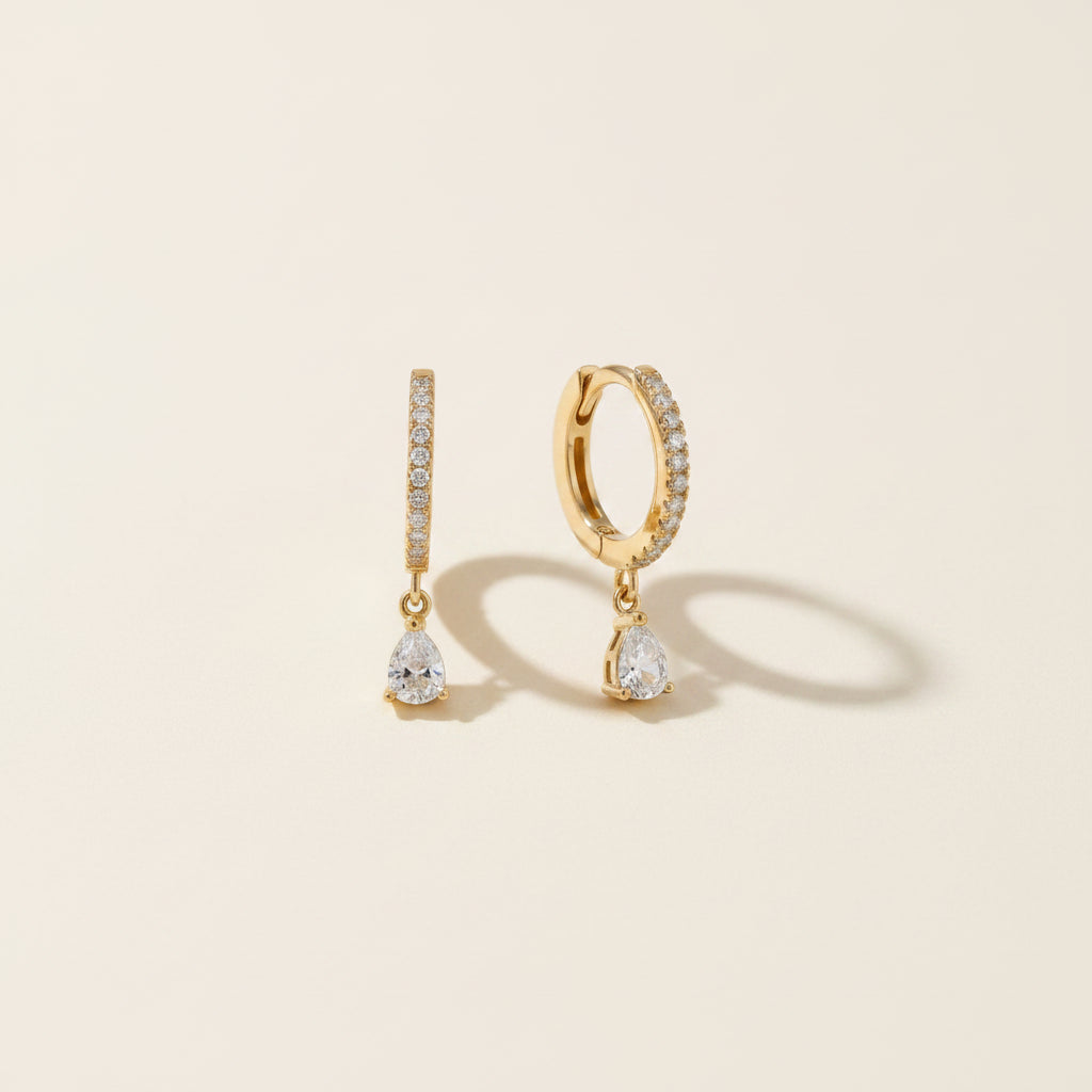A pair of gold huggie hoop earrings with a vertical pavé band and a dangling pear-cut clear gemstone, displayed on a textured beige linen fabric.