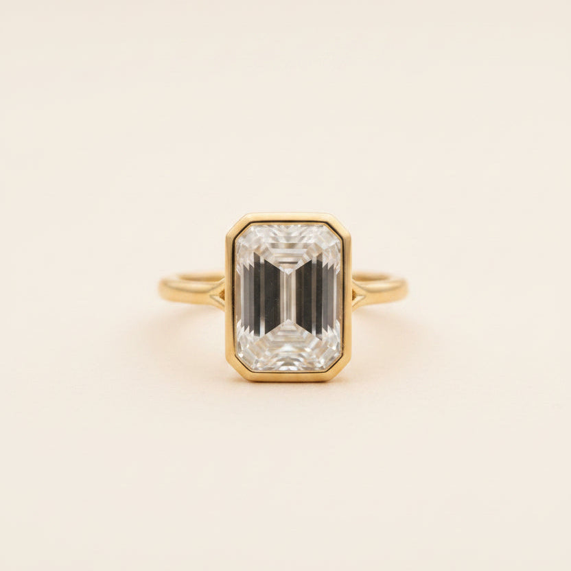 A yellow gold ring with an emerald-cut clear gemstone, resting on a smooth gray stone outdoors with a blurred rocky background.