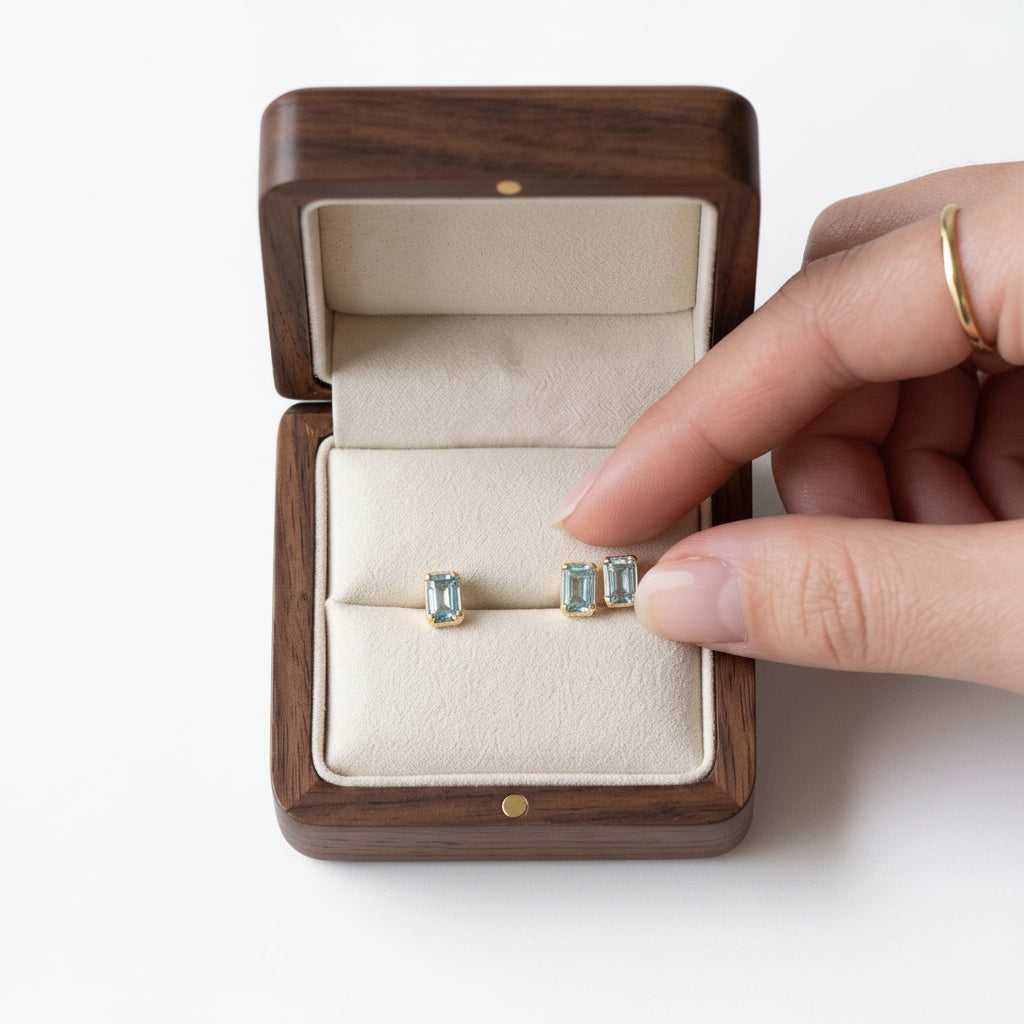 A pair of small stud earrings with light blue rectangular gemstones in silver settings, displayed in an open dark wood jewelry box on a marble surface, with a hand reaching toward them.