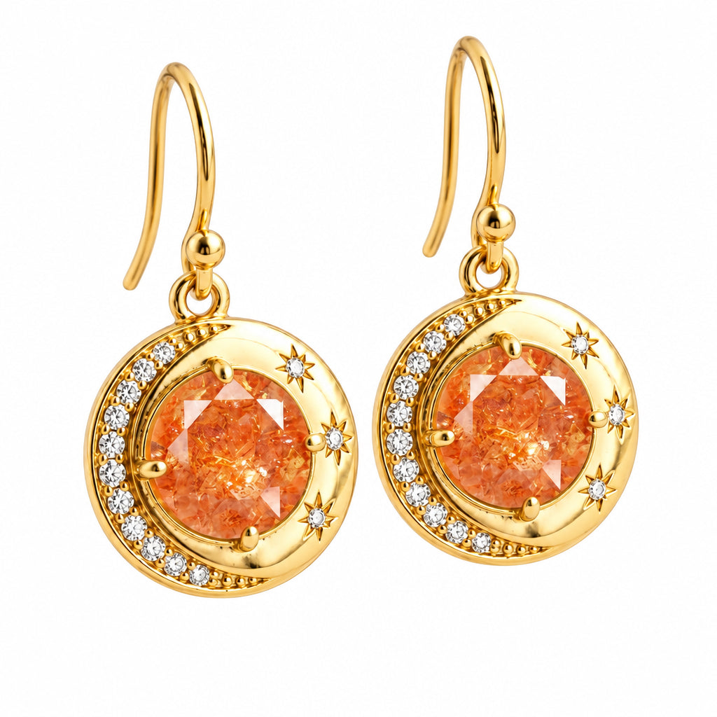 A pair of gold dangle earrings with round orange gemstones, surrounded by a crescent halo of small clear stones and star details, shown against a warm beige bokeh background.