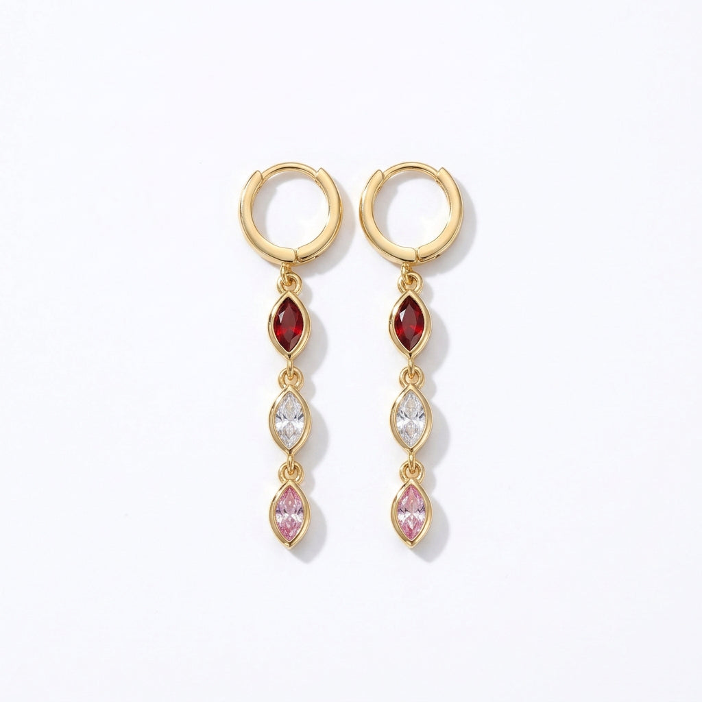 A pair of gold drop earrings with red, clear, and pink marquise-cut stones, displayed on a folded cream linen cloth over a textured stone surface.