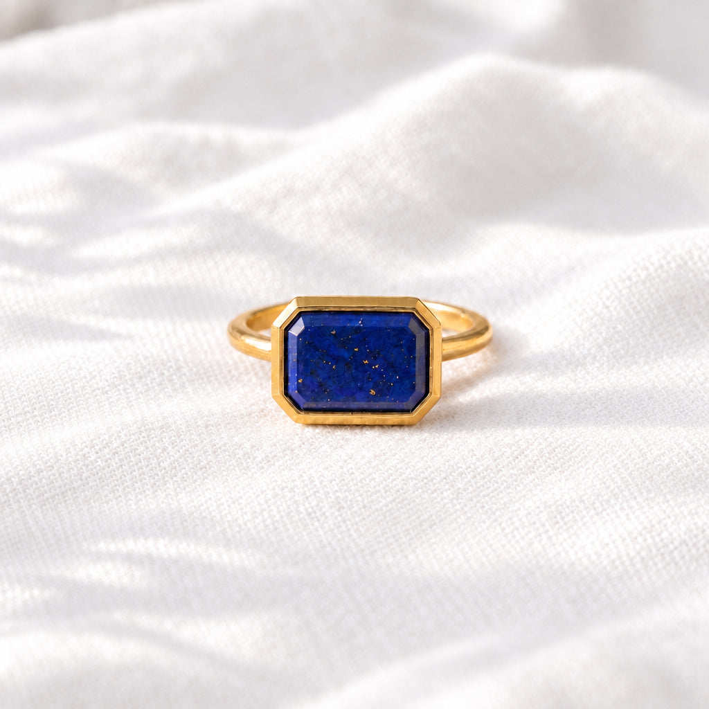 A gold ring with a rectangular blue lapis lazuli stone, resting on a textured cream fabric with dried flowers.