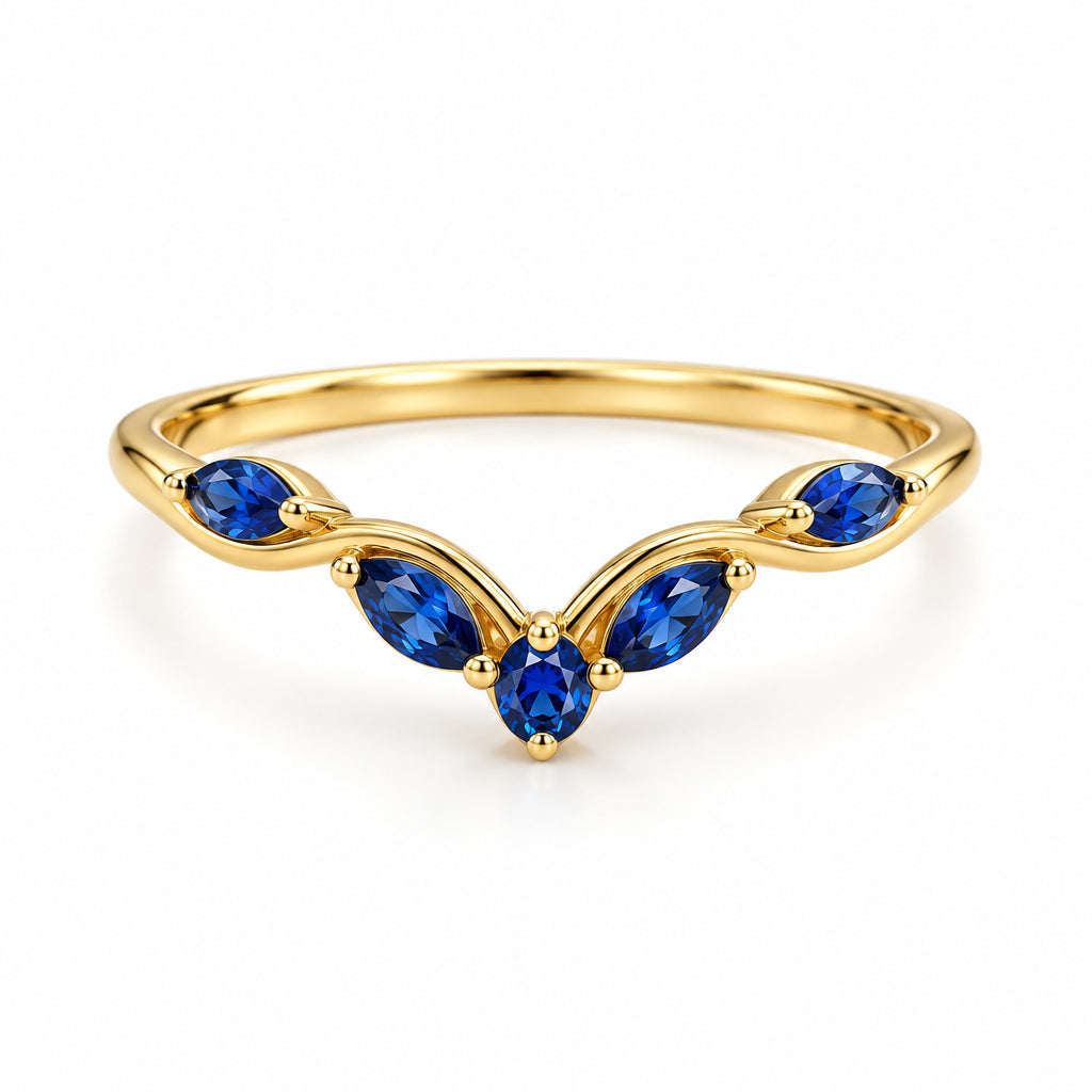 A gold ring with five blue gemstones arranged in a curved, leaf-like pattern, resting on a textured beige fabric with soft green leaves blurred in the background.