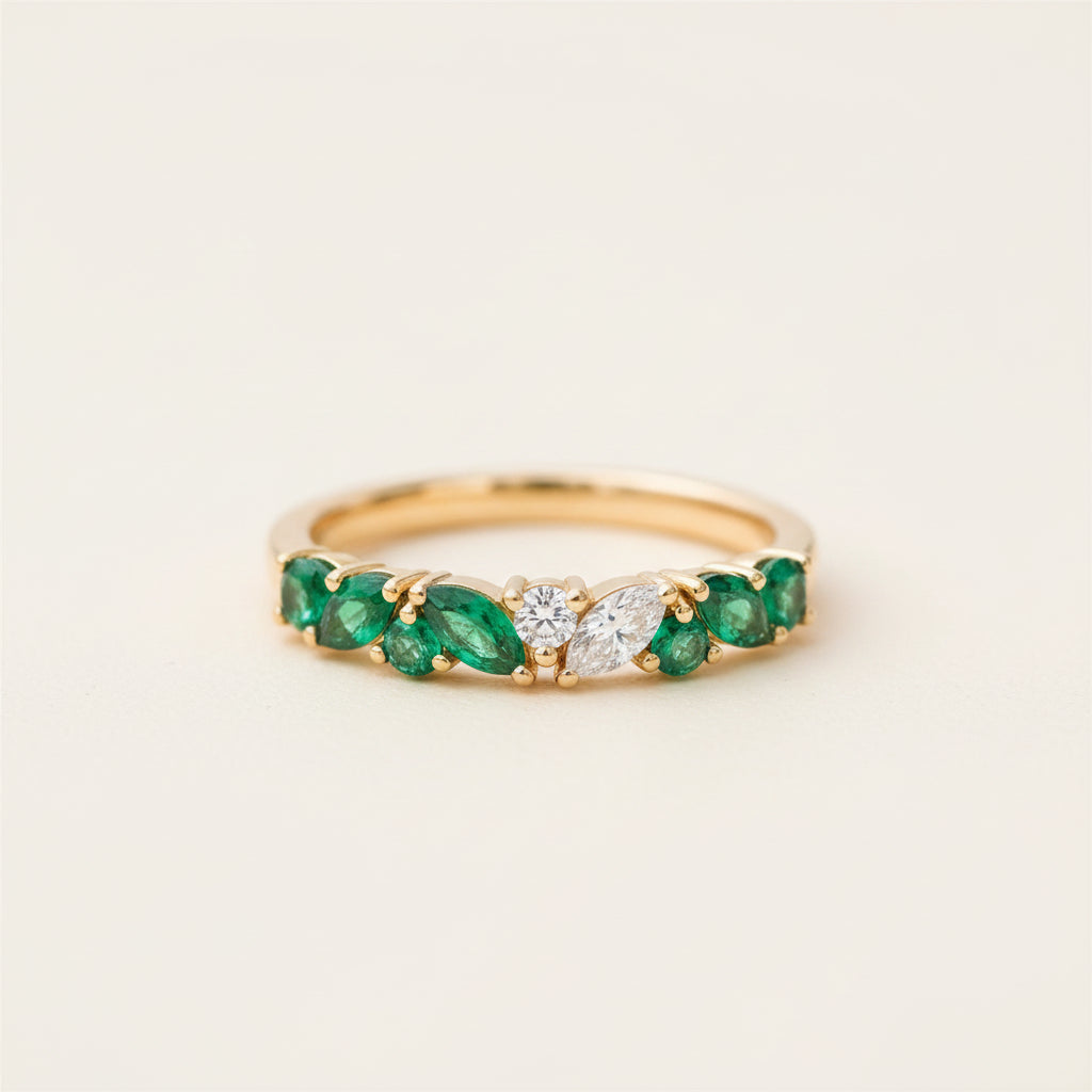 A gold ring featuring a row of green emeralds and a central clear diamond, resting on a textured stone surface with a blurred natural background.