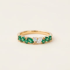 A gold ring featuring a row of green emeralds and a central clear diamond, resting on a textured stone surface with a blurred natural background.