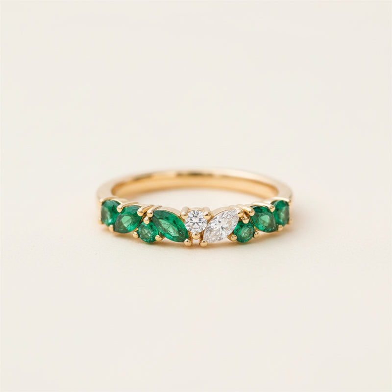 A gold ring featuring a row of green emeralds and a central clear diamond, resting on a textured stone surface with a blurred natural background.