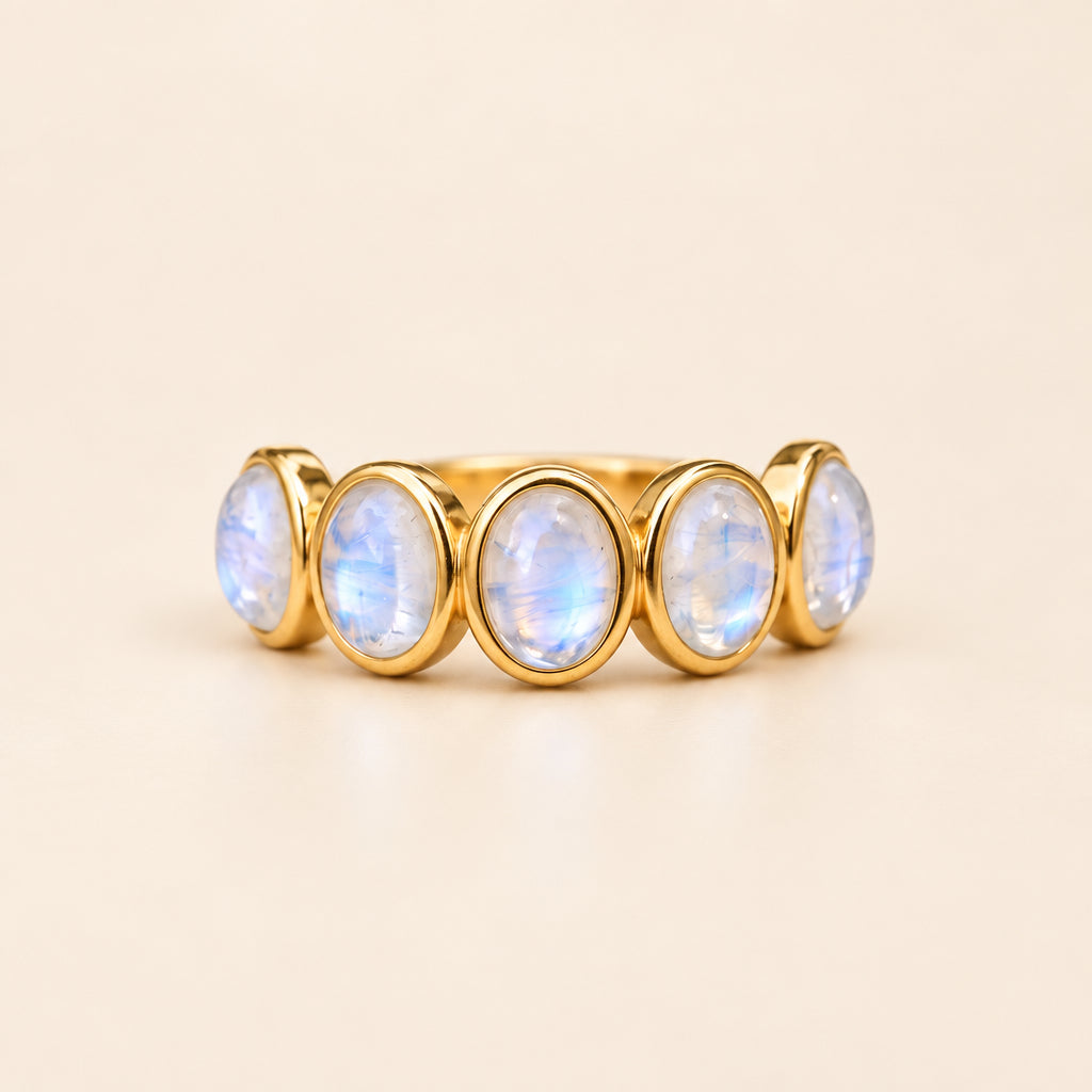 A gold band ring featuring five oval-shaped light blue moonstone gemstones, displayed on a textured beige linen cloth with dried lavender and a pearl in the background.