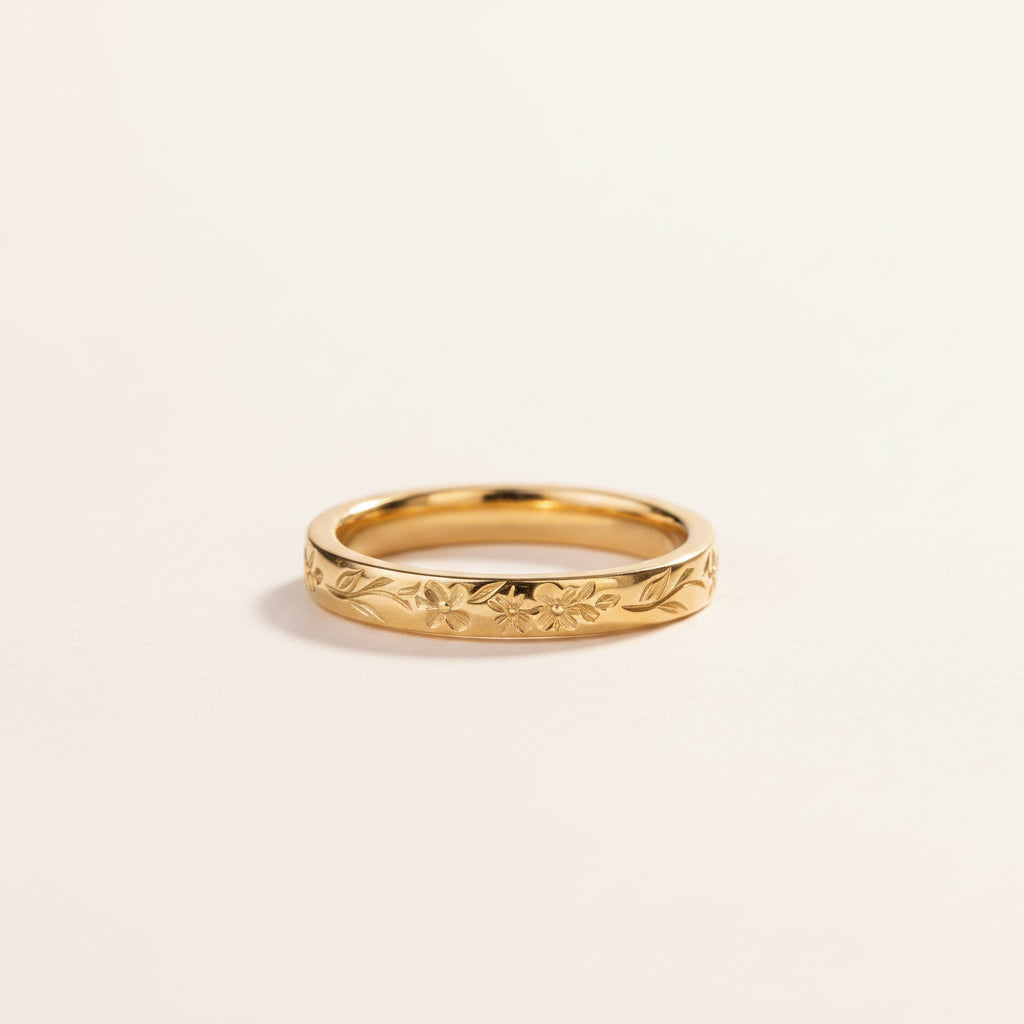 A gold band ring with an engraved floral pattern, resting on a textured stone surface with a soft-focus background of green leaves.
