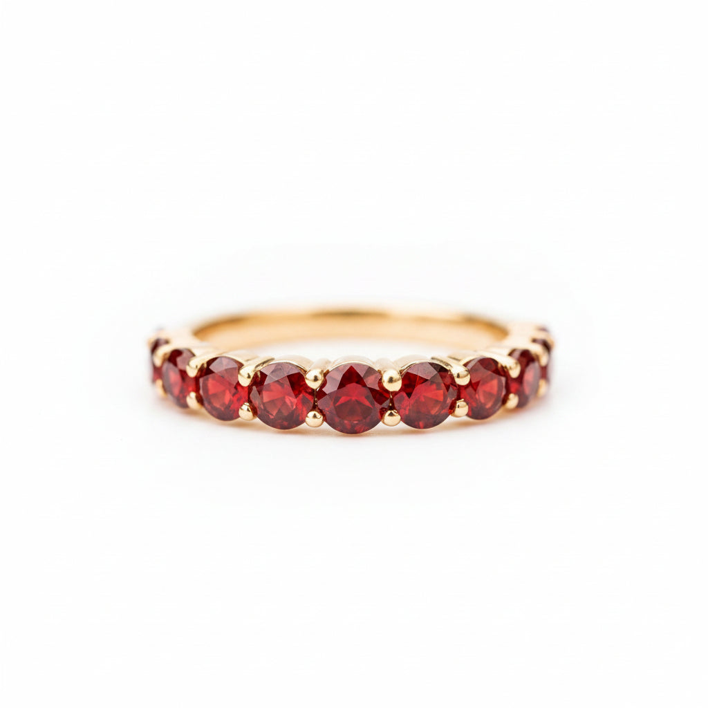 A gold eternity ring featuring a row of round-cut deep red gemstones, resting on a textured stone surface with a soft-focus natural background.