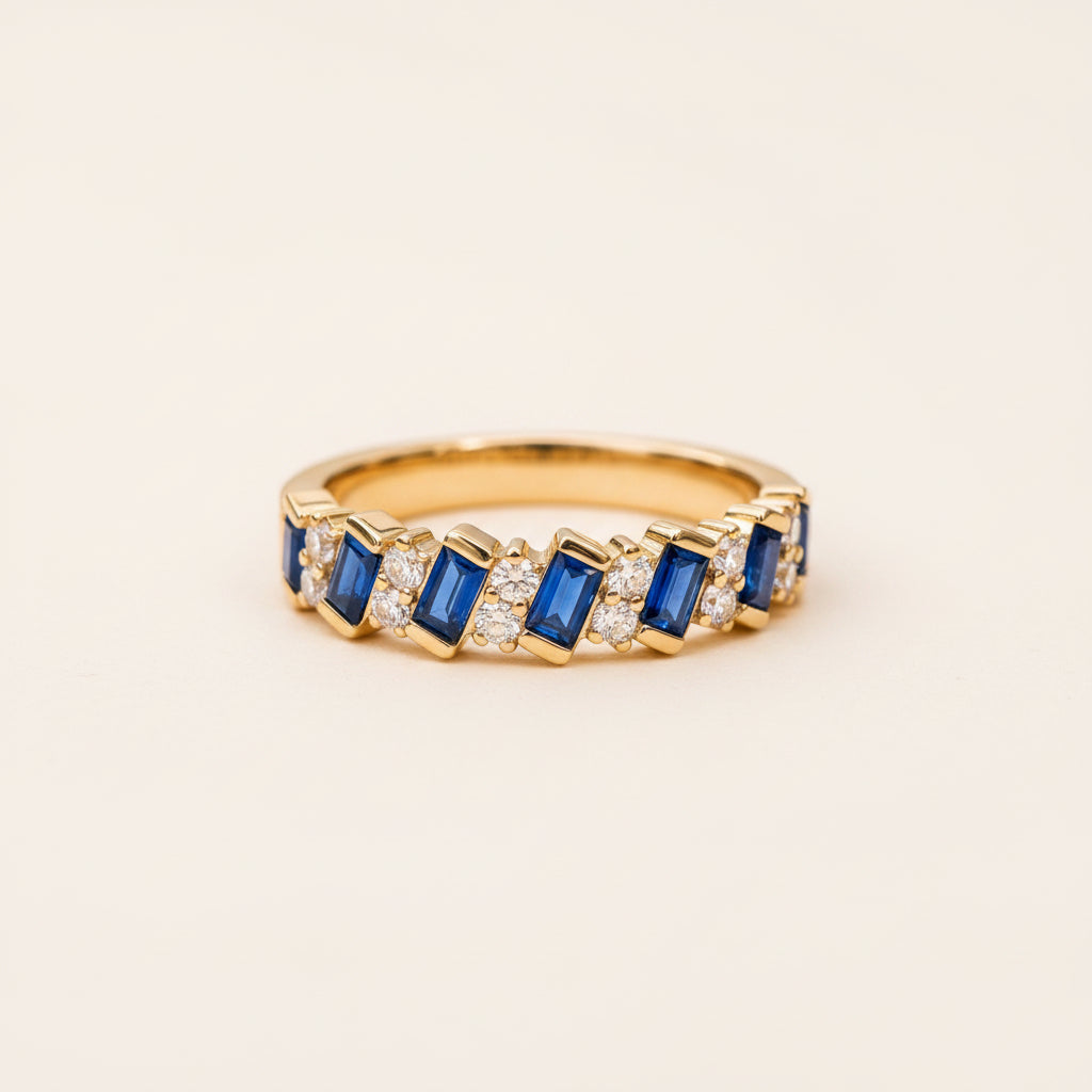 A yellow gold eternity ring featuring baguette-cut blue sapphires and round-cut clear stones, resting on a textured stone surface with a blurred natural background.