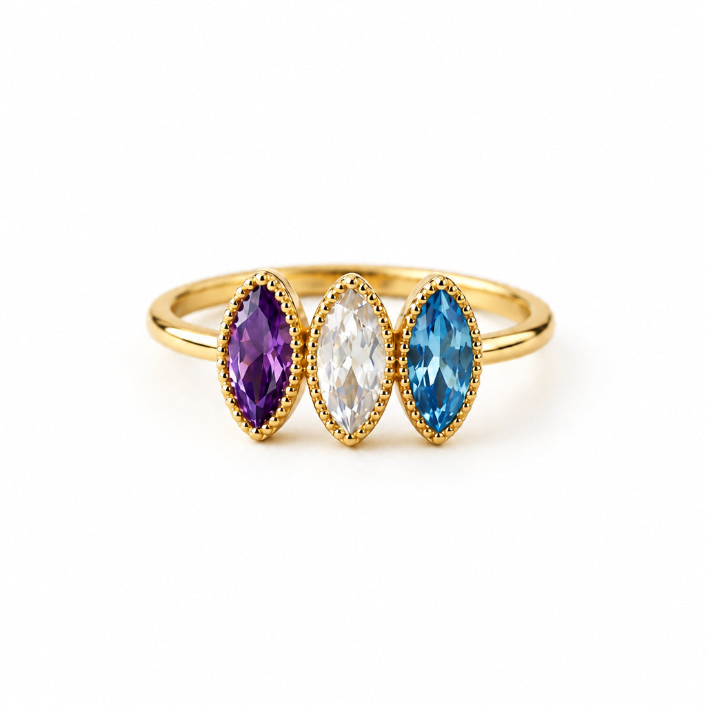 A gold ring with three marquise-cut gemstones in purple, clear, and blue settings, displayed on a textured cream fabric with lace and dried flowers.
