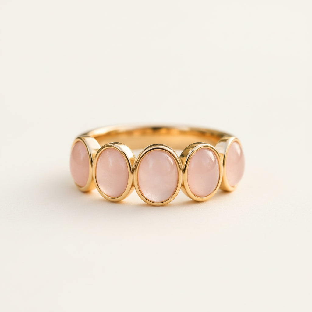 A gold band ring featuring five oval pink gemstones, resting on a textured stone surface with a soft-focus natural background.
