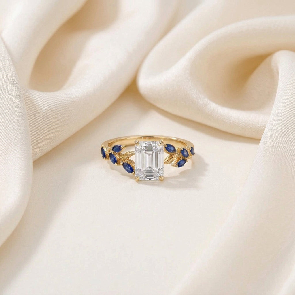 A gold ring with a large emerald-cut clear center stone and blue gemstone accents on the band, styled on a beige stone slab with draped cream fabric and a wooden jewelry box in the background.