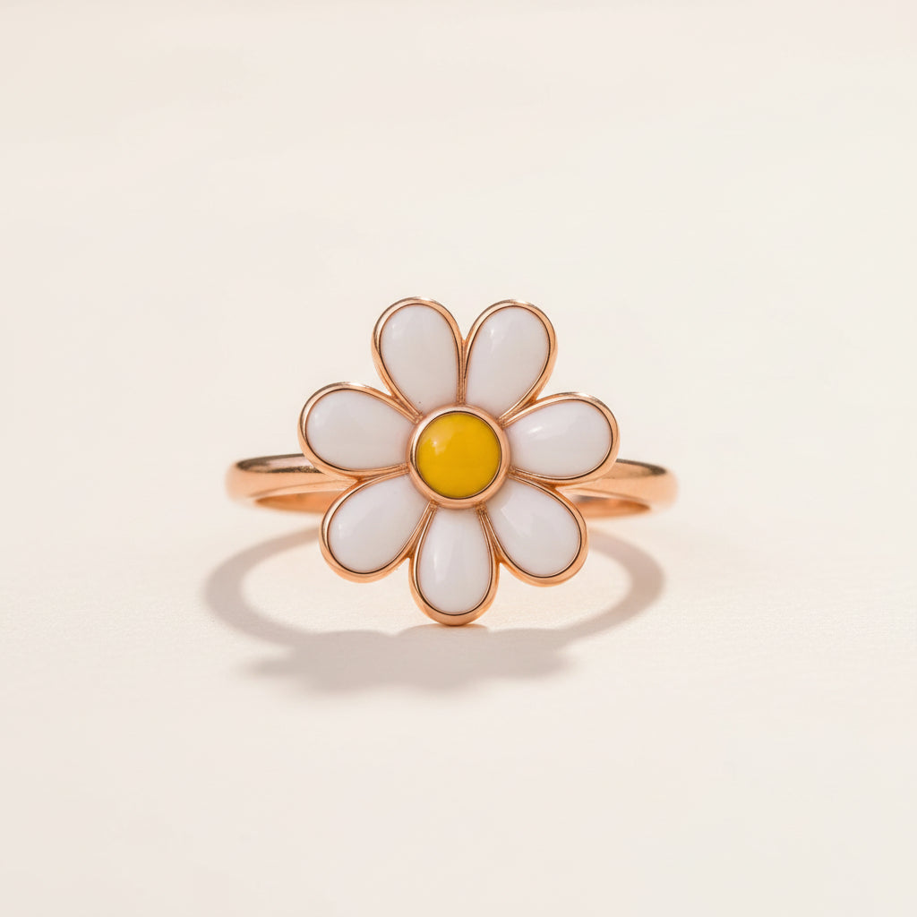 A daisy flower ring with white enamel petals, a yellow center, and a rose gold band, resting on a textured stone with a blurred green background.