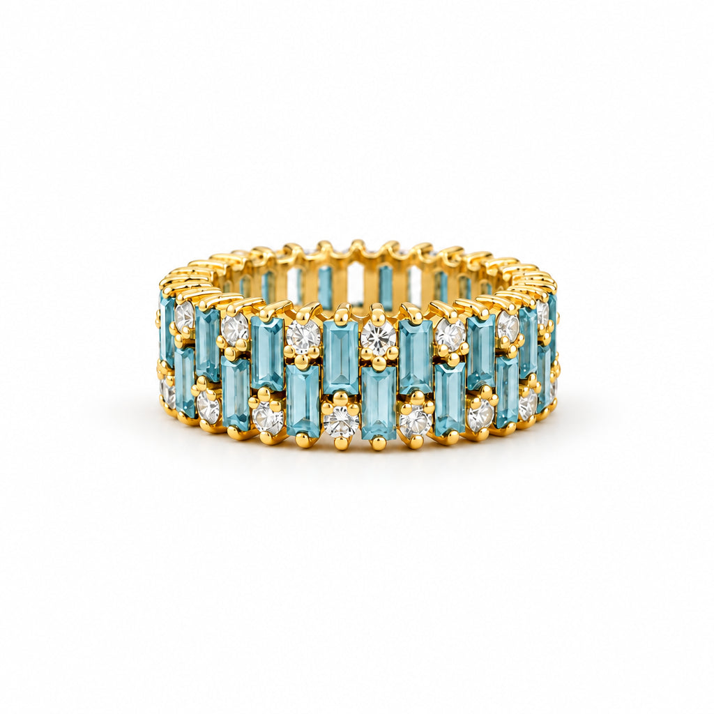 A gold eternity ring featuring light blue baguette and round-cut stones, displayed on a textured beige lace fabric with dried flowers.