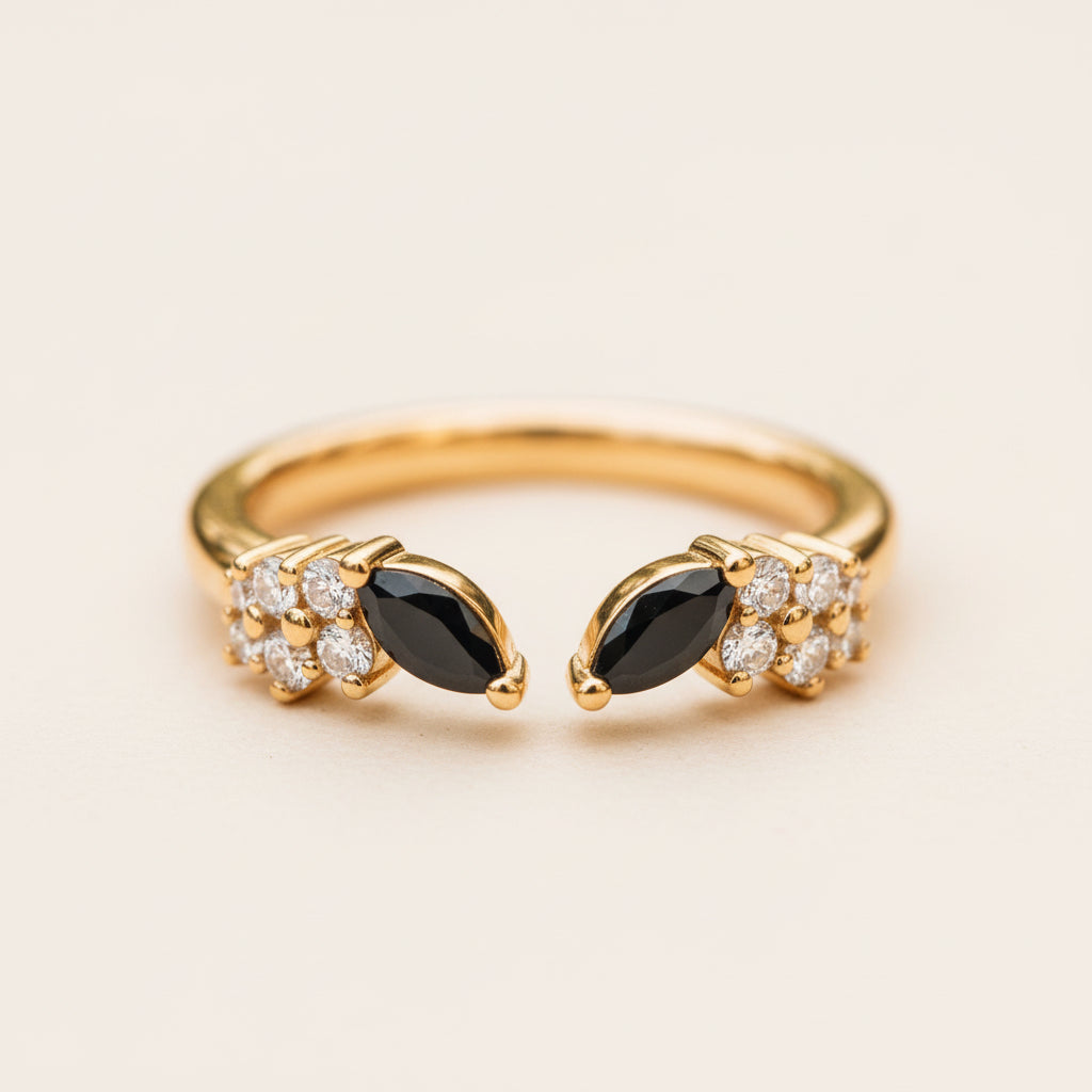 A gold ring with two marquise-cut black onyx centers and clusters of small clear stones, photographed close up on a textured wood surface with a blurred natural background.